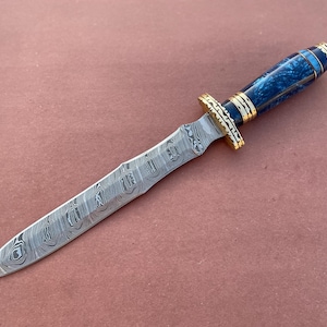 May include: A blue and gold patterned dagger with a long, thin blade. The blade is made of Damascus steel and has a wavy pattern.