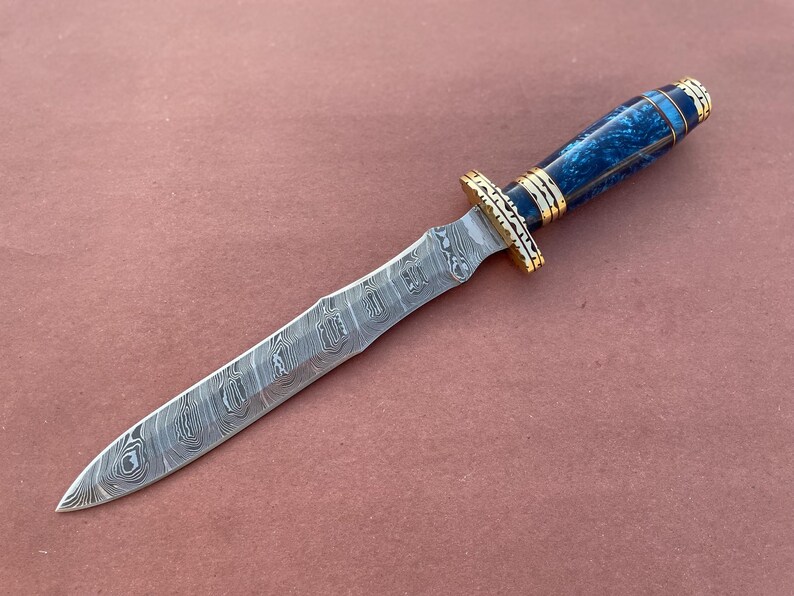 May include: A blue and gold patterned dagger with a long, thin blade. The blade is made of Damascus steel and has a wavy pattern.