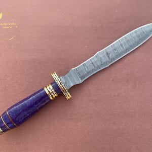 May include: A handcrafted dagger with a patterned steel blade and a purple handle. The handle is accented with gold-colored bands. The blade is approximately 8 inches long. The dagger is displayed on a pinkish-brown surface.