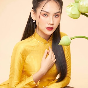 May include: A woman wearing a yellow silk dress with pearl accents sits at a wooden table with a brown teapot and teacups. A green lotus flower is in the background.