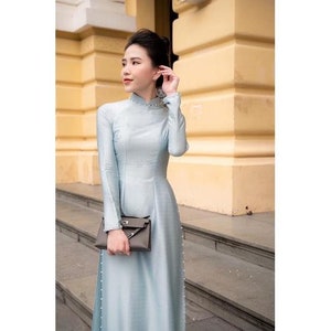 May include: A pale blue, long-sleeved dress with a high neckline and pearl detailing. The dress is worn with a grey clutch. The background shows a building with yellow walls and grey steps.
