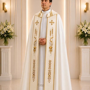 May include: A white liturgical cope featuring gold embroidery of crosses, wheat stalks, and grape clusters along the front panels. The garment is worn over a white cassock with a clerical collar, creating a formal, layered religious vestment.