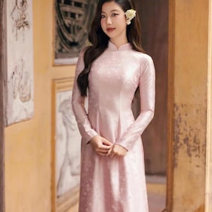 May include: A pale pink, long-sleeved dress with a high collar and a floral pattern. The dress is floor-length and made of a silky material. A flower is placed in the hair.