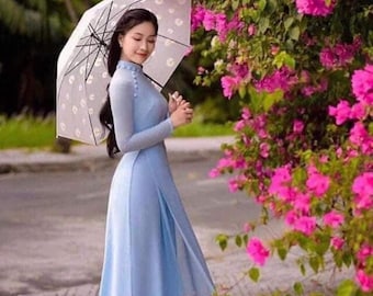 May include: A woman in a light blue dress and holding a white umbrella with a polka dot pattern. The dress has long sleeves and a high collar. Pink flowers and green foliage are in the background.