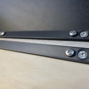 May include: Two black plastic strips with silver screws, each strip has two screws. The strips are on a light wood surface.