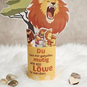 May include: A cylindrical yellow paper container featuring the German text 'Du hast mir geholfen, mutig wie ein Löwe zu werden'. The container holds several paper cutouts of lions and a tree, including a large lion with an open mouth.