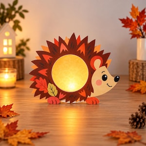 Op de afbeelding: Een decoratieve egelvormige lamp met een cirkelvormig gloeiend midden, bruine en oranje stekels met bladpatroon en een lichtgekleurd gezicht met een kleine zwarte neus en oog. De lamp verspreidt een warm geel licht vanuit de kern.