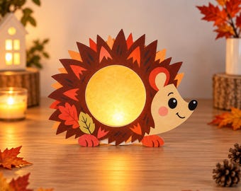 Puede incluir: Una lámpara decorativa con forma de erizo que presenta un centro circular brillante, púas de color marrón y naranja, y una cara clara con una pequeña nariz y un ojo negros. El diseño incorpora motivos de hojas de otoño en tonos rojos, naranjas y amarillos.