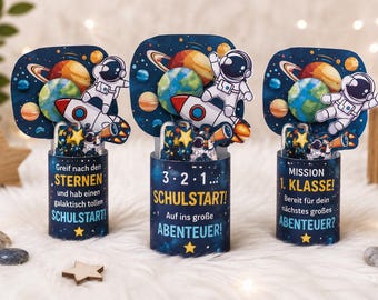 May include: Three cylindrical containers featuring space-themed graphics with astronauts, rockets, and planets. The text reads: Greif nach den STERNEN und hab einen galaktisch tollen SCHULSTART!, 3·2·1... SCHULSTART! Auf ins große ABENTEUER!, and MISSION 1. KLASSE! Bereit für dein nächstes großes ABENTEUER?