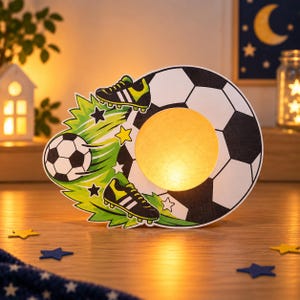 Könnte beinhalten: Ein dekoratives Nachtlicht im Fußball-Design mit einer großen schwarz-weißen Fußballgrafik, zwei schwarzen und neongrünen Fußballschuhen und einem leuchtenden kreisförmigen Zentrum. Das Design enthält grüne Grasakzente und kleine Sterne.