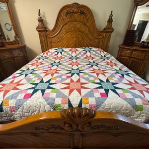 May include: A bed quilt featuring a repeating star pattern in shades of coral, teal, pink, yellow, and light blue on a white background. The quilt has intricate, swirling machine-stitched quilting patterns throughout the entire fabric surface.