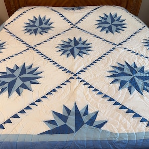 May include: A white quilt featuring a repeating pattern of blue mariner's compass stars in various shades of blue. The quilt is constructed with diamond-shaped blocks separated by blue sawtooth borders and finished with intricate curved quilting stitches.