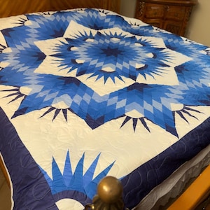 May include: A quilt featuring a large, central starburst pattern in shades of navy, royal, and light blue against a white background. The design consists of geometric diamond shapes and radiating points, finished with a solid dark blue border.