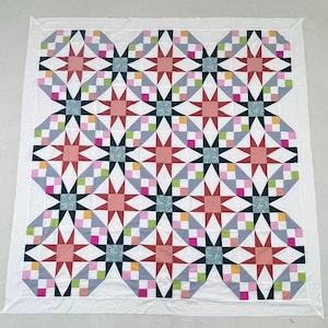 May include: A square quilt featuring a repeating star pattern in shades of red, pink, and dark blue, accented with small multi-colored checkered squares in green, yellow, and pink, all set against a white background with a solid white border.