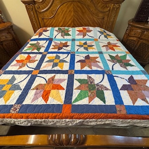 May include: A quilt featuring a repeating pattern of multi-colored fabric leaves in square blocks. The leaves are constructed from various patterned and solid fabrics in shades of orange, yellow, green, blue, and brown, separated by blue and white sashing.