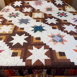 May include: A patchwork quilt featuring a repeating star pattern in white, brown, and tan fabric. The design incorporates various floral and geometric printed fabrics within the star centers, set against a background of white and brown blocks.