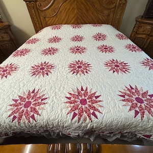May include: A white bed quilt featuring a repeating pattern of large, multi-pointed star motifs in shades of pink and red. The fabric surface displays intricate, swirling quilted stitching throughout, creating a textured appearance across the entire cover.