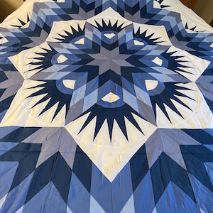 May include: A large quilt featuring a central star pattern in shades of navy, royal, and light blue, accented with white geometric shapes. The design incorporates radiating points and diamond-shaped fabric segments arranged in a symmetrical layout.