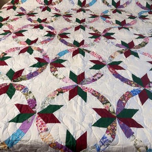 May include: A quilt featuring a repeating pattern of interlocking circles and star shapes. The design incorporates a white background with fabric segments in floral prints, solid dark green, and solid maroon, all joined by intricate stitching.