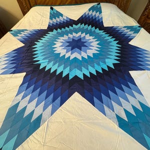 May include: A quilt featuring a large central eight-pointed star pattern composed of diamond-shaped fabric pieces in various shades of blue, including navy, royal, and light blue, set against a solid white background covering the entire surface.