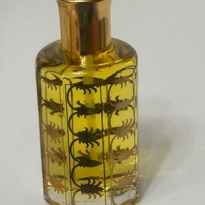 May include: A small, ornate perfume bottle with a gold-colored cap and a red gem on top. The clear glass bottle is filled with a yellow liquid and decorated with a gold leaf pattern. The bottle has a vintage or antique aesthetic.