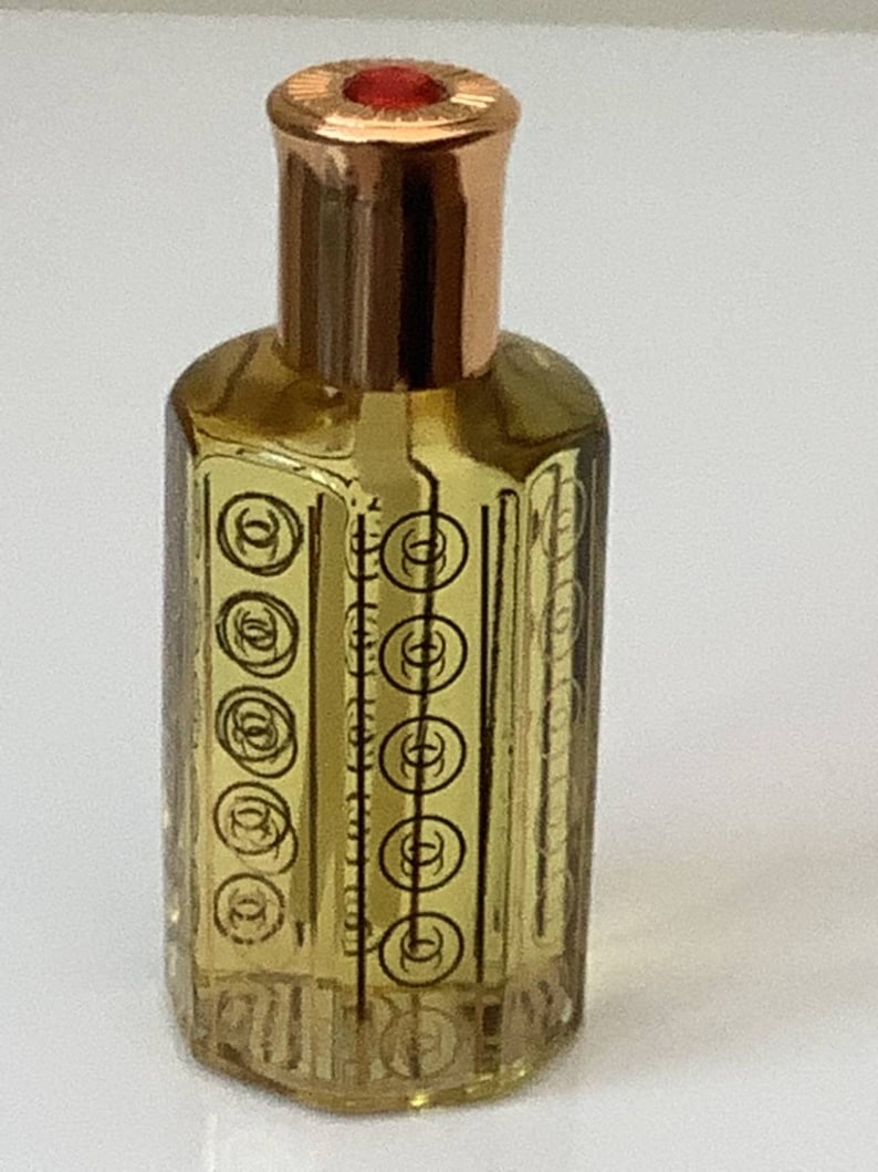 May include: A small, clear glass bottle with a gold cap and a red top. The bottle has a decorative pattern of circles and lines.