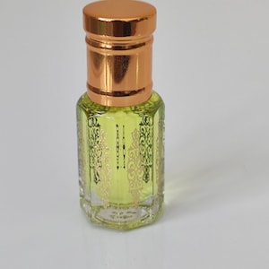 May include: A small, clear glass perfume bottle with a gold-coloured cap and decorative gold filigree. The bottle contains a light yellow liquid. The cap and the bottle's top section are a shiny gold colour.