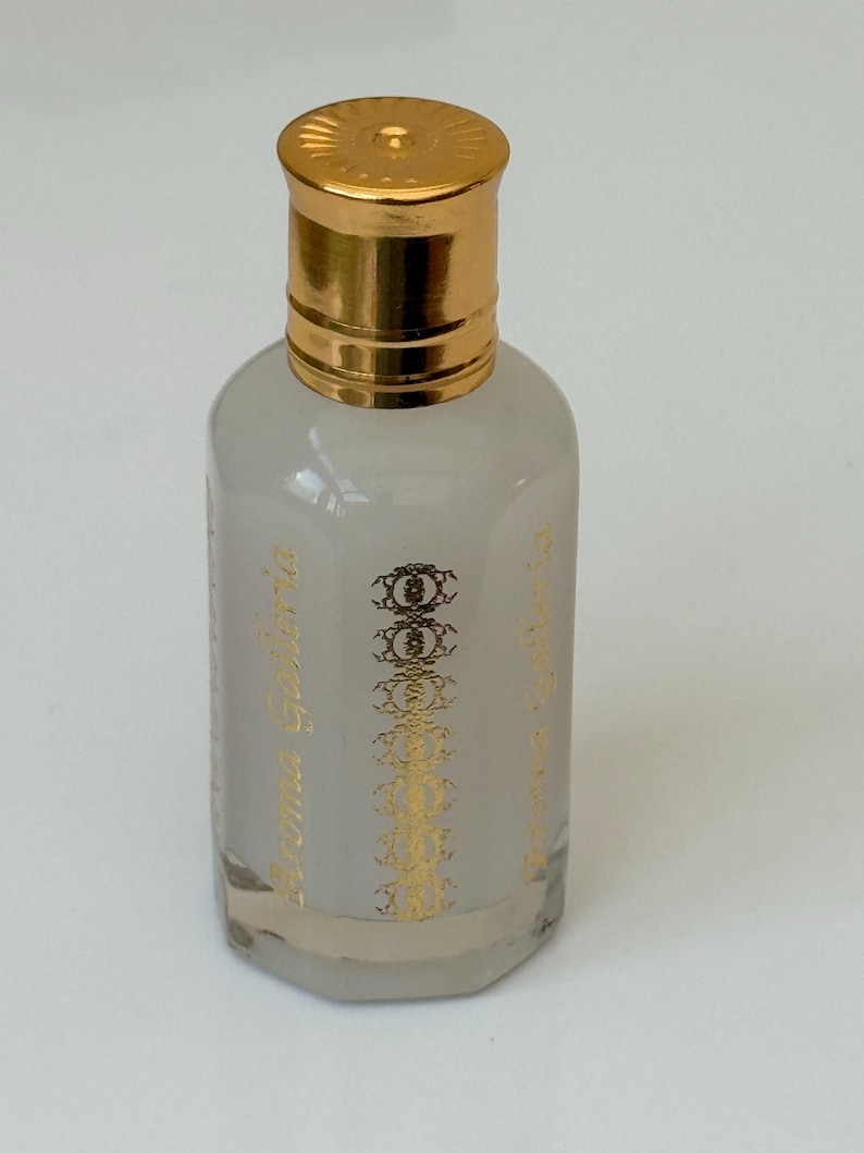 Pode incluir: Uma pequena garrafa de vidro transparente com tampa dourada. A garrafa cont&eacute;m um l&iacute;quido cinza claro e texto e elementos decorativos dourados. O texto na garrafa diz "Aroma Galleria".