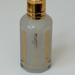 Pode incluir: Frasco de perfume pequeno e transparente com tampa dourada. O frasco tem um tom cinza claro e &eacute; decorado com desenhos dourados e o texto "Aroma Galleria". O frasco est&aacute; cheio de um l&iacute;quido transparente.