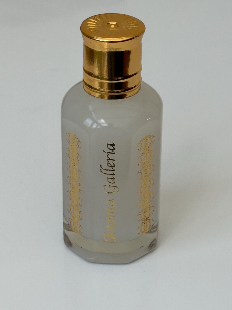 Pode incluir: Frasco de perfume pequeno e transparente com tampa dourada. O frasco tem um tom cinza claro e &eacute; decorado com desenhos dourados e o texto "Aroma Galleria". O frasco est&aacute; cheio de um l&iacute;quido transparente.