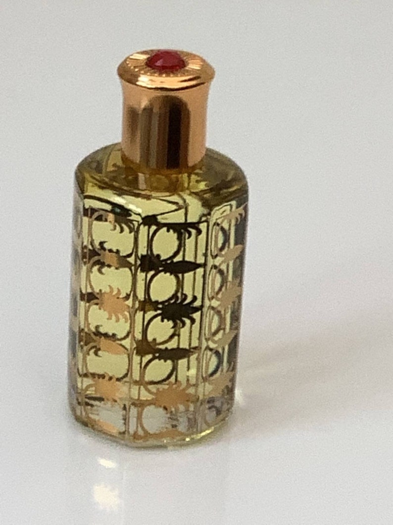May include: A small, clear glass bottle with a gold cap and a red top. The bottle is filled with a light yellow liquid and has a gold patterned design.