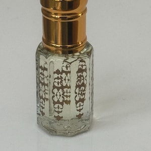 Könnte beinhalten: Eine kleine, durchsichtige Glasflasche mit einem goldenen Verschluss. Die Flasche hat ein dekoratives Muster an den Seiten und ist mit einer klaren Flüssigkeit gefüllt.