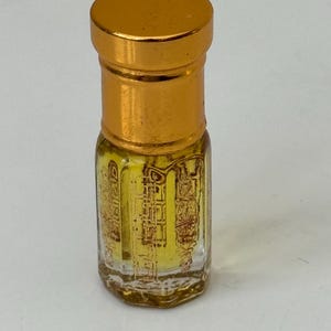 May include: A small, clear glass perfume bottle with a gold-colored cap and a gold-colored base. The bottle contains a yellow liquid and has gold-colored decorative detailing on the glass. The bottle is standing upright.