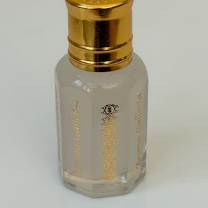 Pode incluir: Frasco de perfume pequeno, transparente, com tampa dourada. O frasco tem um r&oacute;tulo dourado com o texto "Aroma Galleria" e um design ornamental. O frasco est&aacute; cheio de um l&iacute;quido claro.