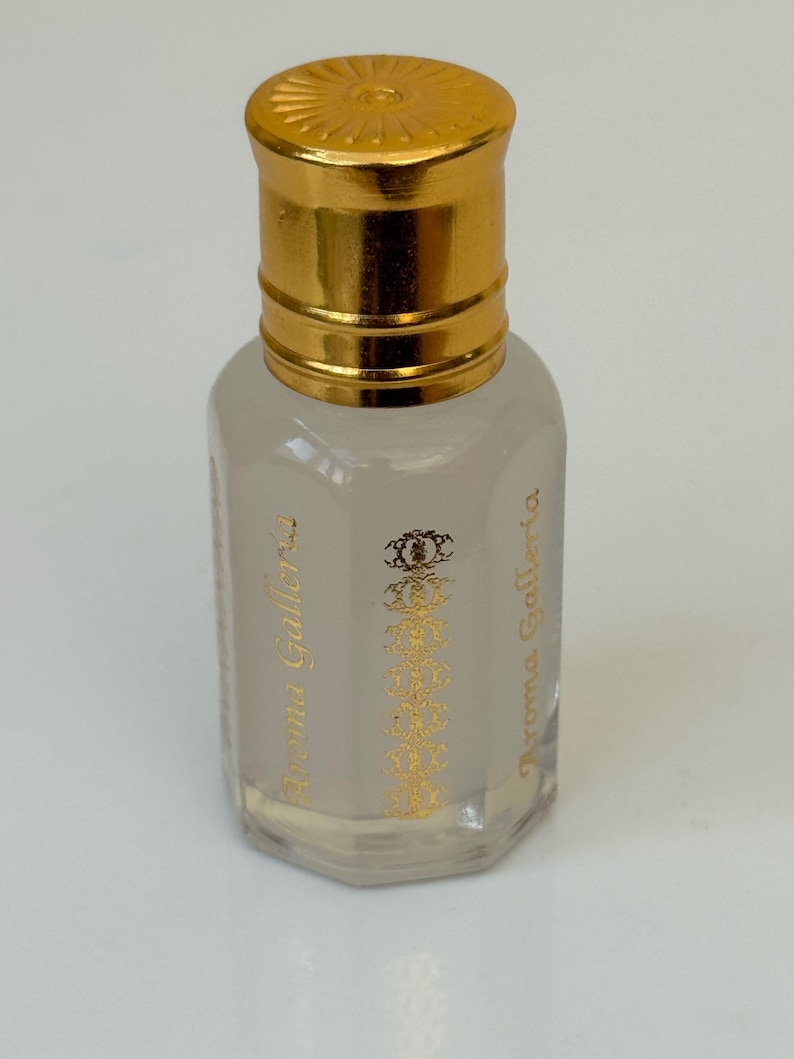 Pode incluir: Frasco de perfume pequeno, transparente, com tampa dourada. O frasco tem um r&oacute;tulo dourado com o texto "Aroma Galleria" e um design ornamental. O frasco est&aacute; cheio de um l&iacute;quido claro.