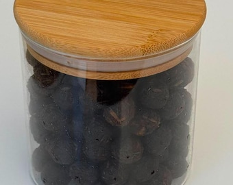 Op de afbeelding: Een cilindrische voorraadpot van helder glas gevuld met donkerbruine, bolvormige, getextureerde objecten. De pot is afgesloten met een rond, lichtgekleurd bamboe deksel met een zichtbare houtnerf en een siliconen afdichtingsring.