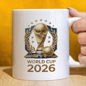May include: A white ceramic mug featuring a gold-colored World Cup trophy graphic centered above a soccer ball. The design includes a blue shield, gold laurel leaves, and stars. The text reads WORLD CUP 2026 in bold, dark-colored lettering.