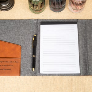 May include: A gray and brown leatherette folder with a pen and a lined notepad. The folder has a pocket on the front with the quote "The future belongs to those who believe in the beauty of their dreams." -Eleanor Roosevelt.
