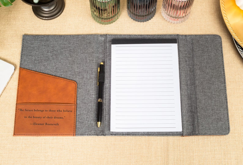 May include: A gray and brown leatherette folder with a pen and a lined notepad. The folder has a pocket on the front with the quote "The future belongs to those who believe in the beauty of their dreams." -Eleanor Roosevelt.