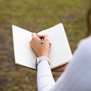 May include: A person writing in a lined notebook with a pen. The notebook is open and the person is holding it with their left hand. The person is wearing a white shirt and a watch on their left wrist.
