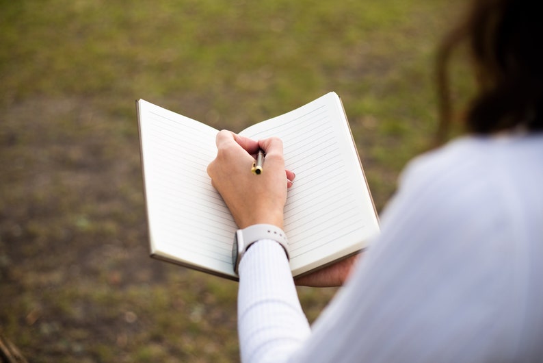 May include: A person writing in a lined notebook with a pen. The notebook is open and the person is holding it with their left hand. The person is wearing a white shirt and a watch on their left wrist.