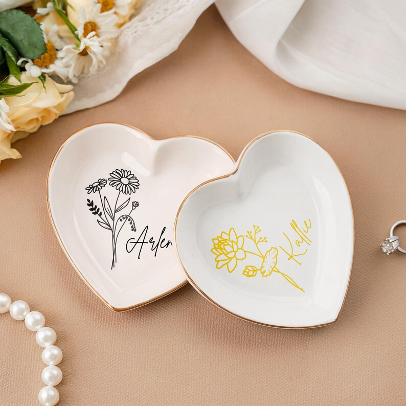 Op de afbeelding: Twee witte hartvormige keramische schalen met gouden rand. De schaal links heeft een zwart bloemenmotief en de naam "Arlen" eronder geschreven. De schaal rechts heeft een geel bloemenmotief en de naam "Kallie" eronder geschreven.