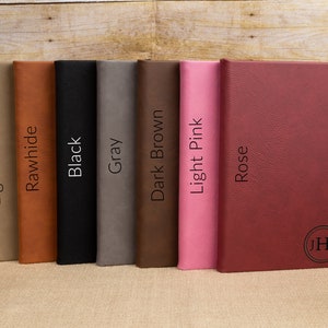 May include: Seven different colored leather-bound journals lined up on a wooden surface. The colors are light brown, tan, black, gray, dark brown, light pink, and rose. The last journal has the initials "JHK" embossed on the cover.