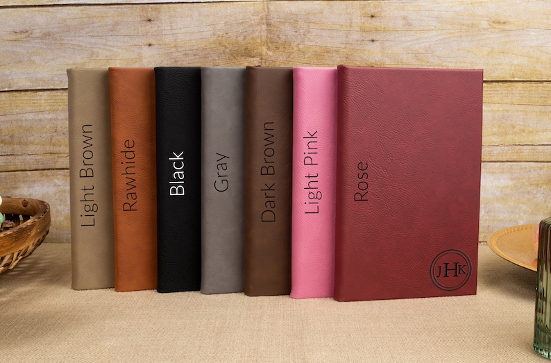 May include: Seven different colored leather-bound journals lined up on a wooden surface. The colors are light brown, tan, black, gray, dark brown, light pink, and rose. The last journal has the initials "JHK" embossed on the cover.