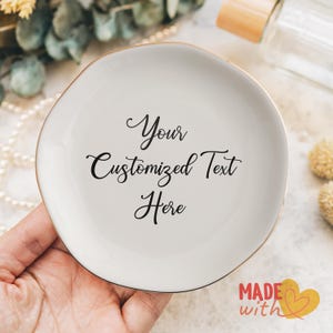 May include: White ceramic ring dish with a gold rim. The dish has the text "Your Customized Text Here" written in black cursive.