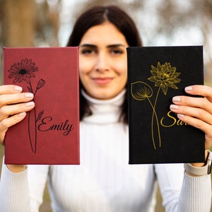 May include: Two personalized journals with floral designs. The journal on the left is red with a black daisy and the name "Emily" written on it. The journal on the right is black with a gold lotus flower and the name "Sarah" written on it.