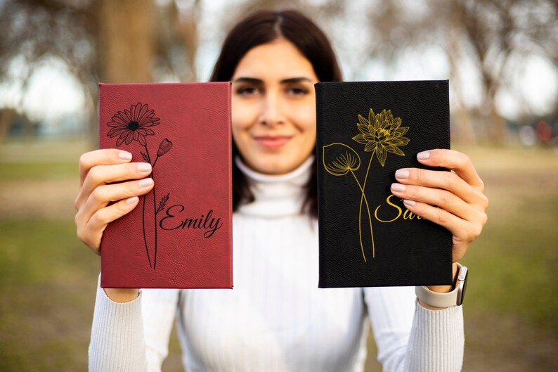 May include: Two personalized journals with floral designs. The journal on the left is red with a black daisy and the name "Emily" written on it. The journal on the right is black with a gold lotus flower and the name "Sarah" written on it.