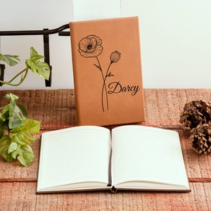 May include: A brown leather journal with a poppy flower design and the name "Darcy" engraved on the front cover. The journal is open to a blank page with lined paper.