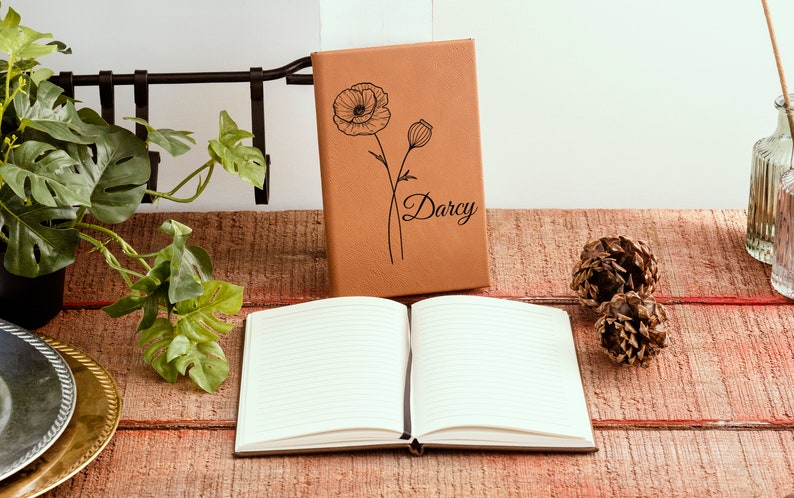 May include: A brown leather journal with a poppy flower design and the name "Darcy" engraved on the front cover. The journal is open to a blank page with lined paper.
