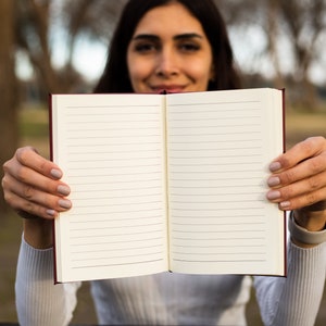 May include: A person holding a blank, lined notebook open with both hands. The notebook has a red cover and white pages.